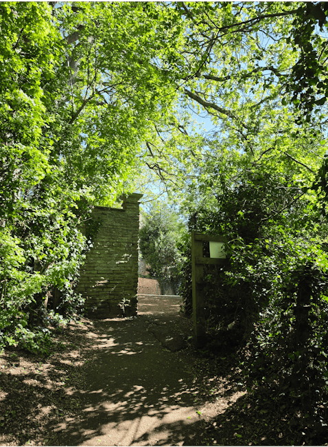 A neatly maintained garden in front of a traditional red brick house with white-framed windows and a steep, tiled roof. The garden features a well-trimmed hedge border surrounding a lush, green lawn a
